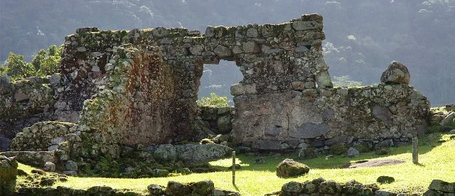 Vista das ruínas do Engenho São Jorge dos Erasmos, com paredes de pedra preservadas entre árvores.